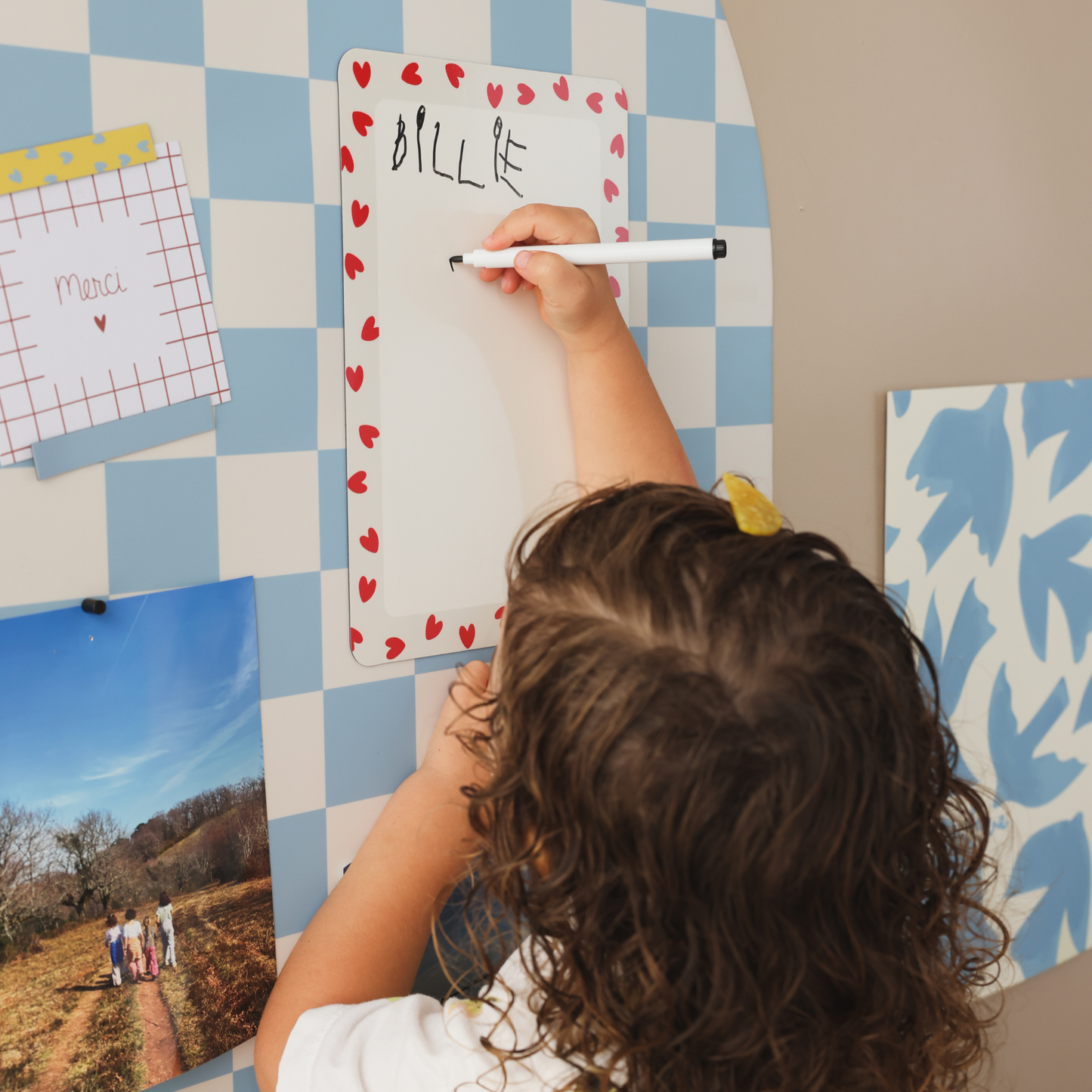 Enfant écrivant son prénom au feutre effaçable à sec noir sur ardoise magnétique rectangle coeurs rouges, aimantée sur tableau magnétique arche damier bleu