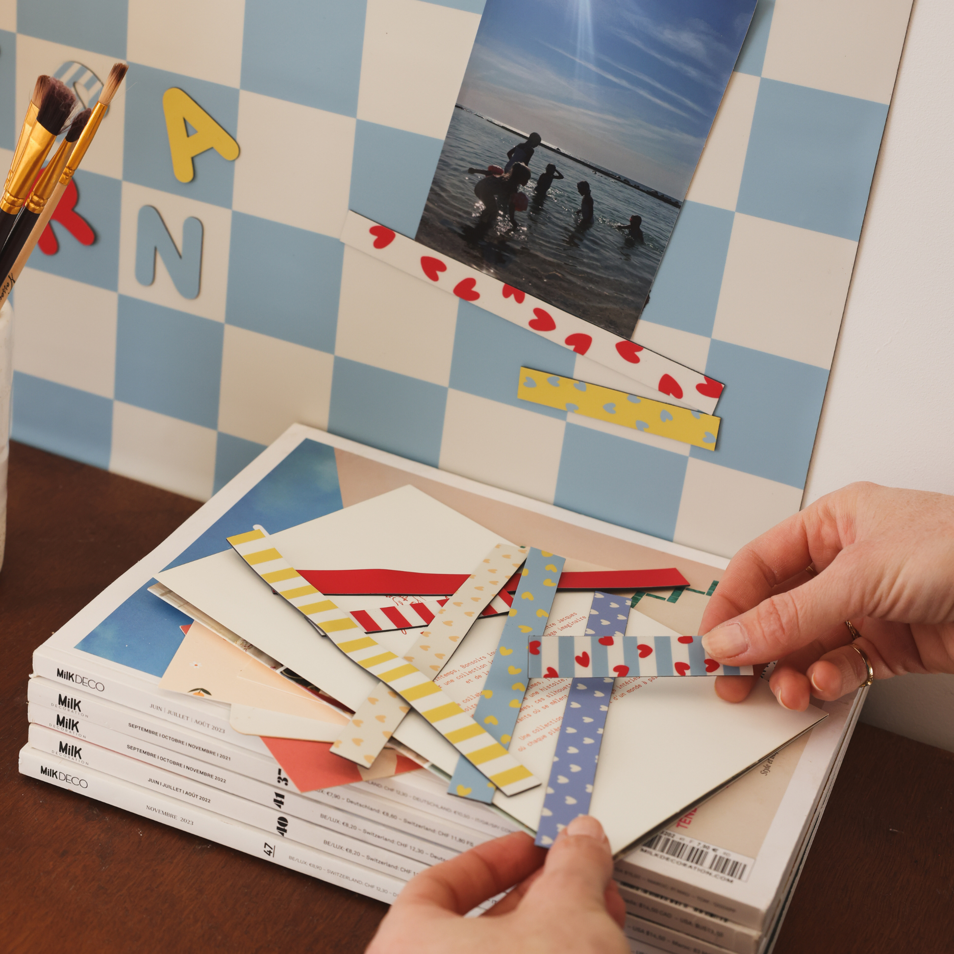 Mains assemblant des bandes de masking tape magnétique colorés sur des magazines à côté d'une photo de vacances sur tableau magnétique damier bleu