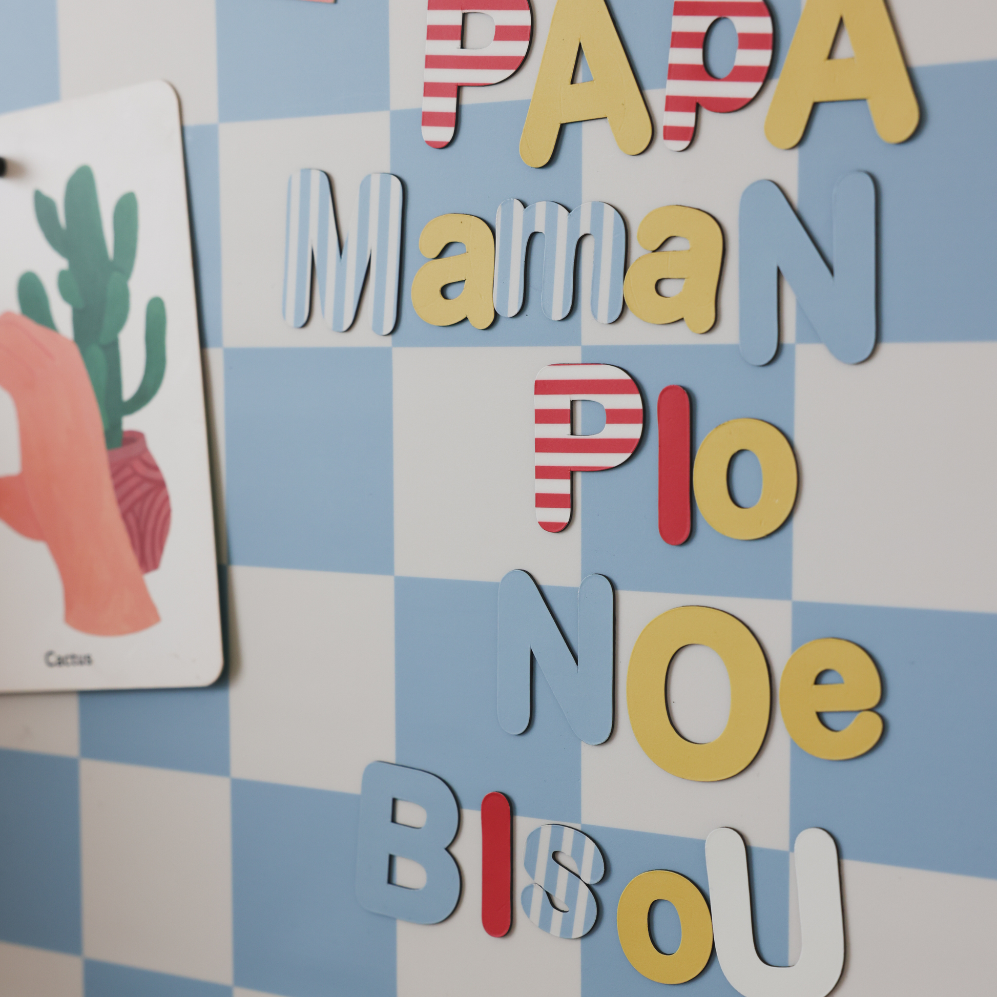 Gros plan sur les lettres magnétiques Primaires composant des mots Papa, Maman, Pio sur support mural damier bleu et beige