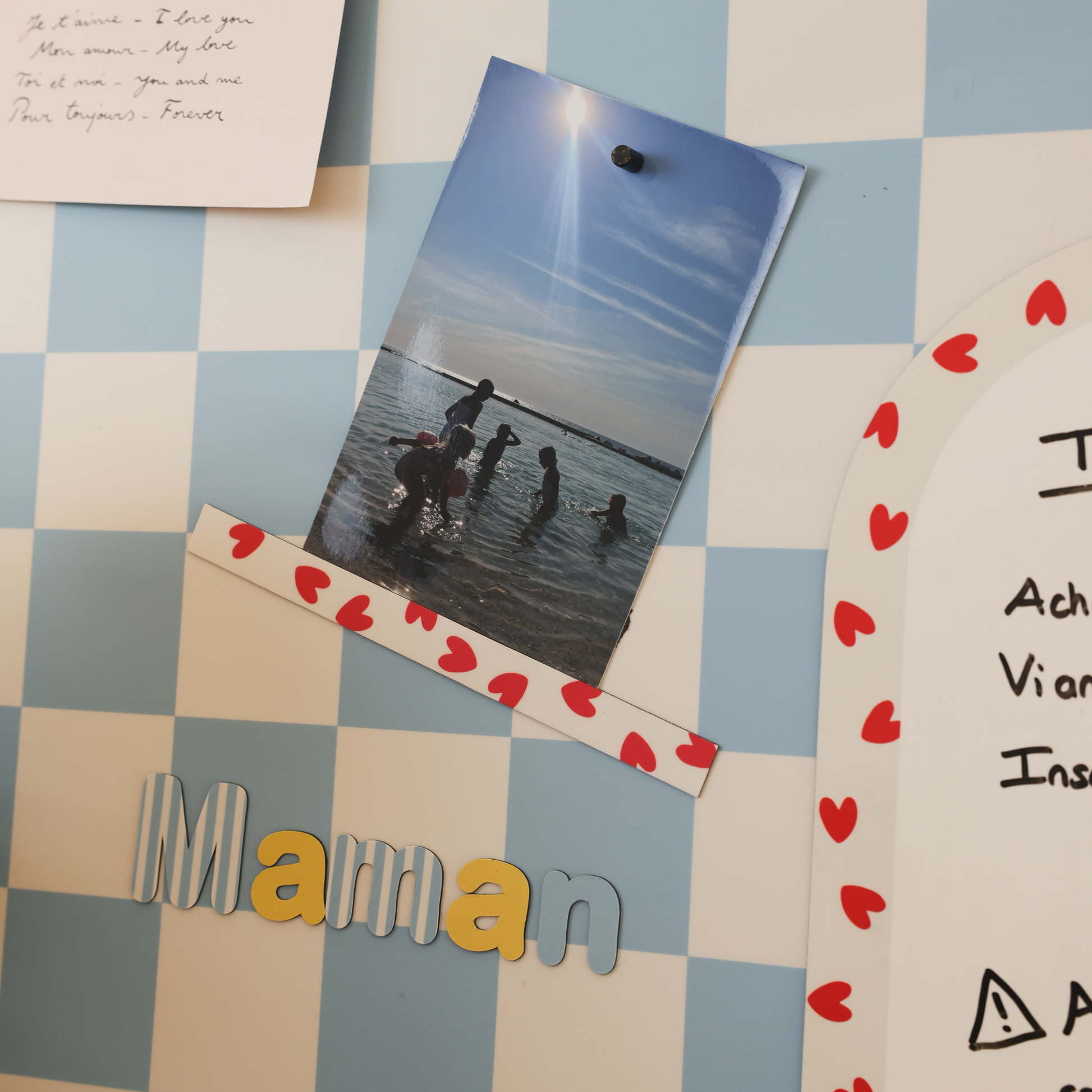 Masking tape magnétique à motif coeurs rouges fixant une photo de bord de mer sur revêtement damier bleu et beige, lettres magnétiques Maman